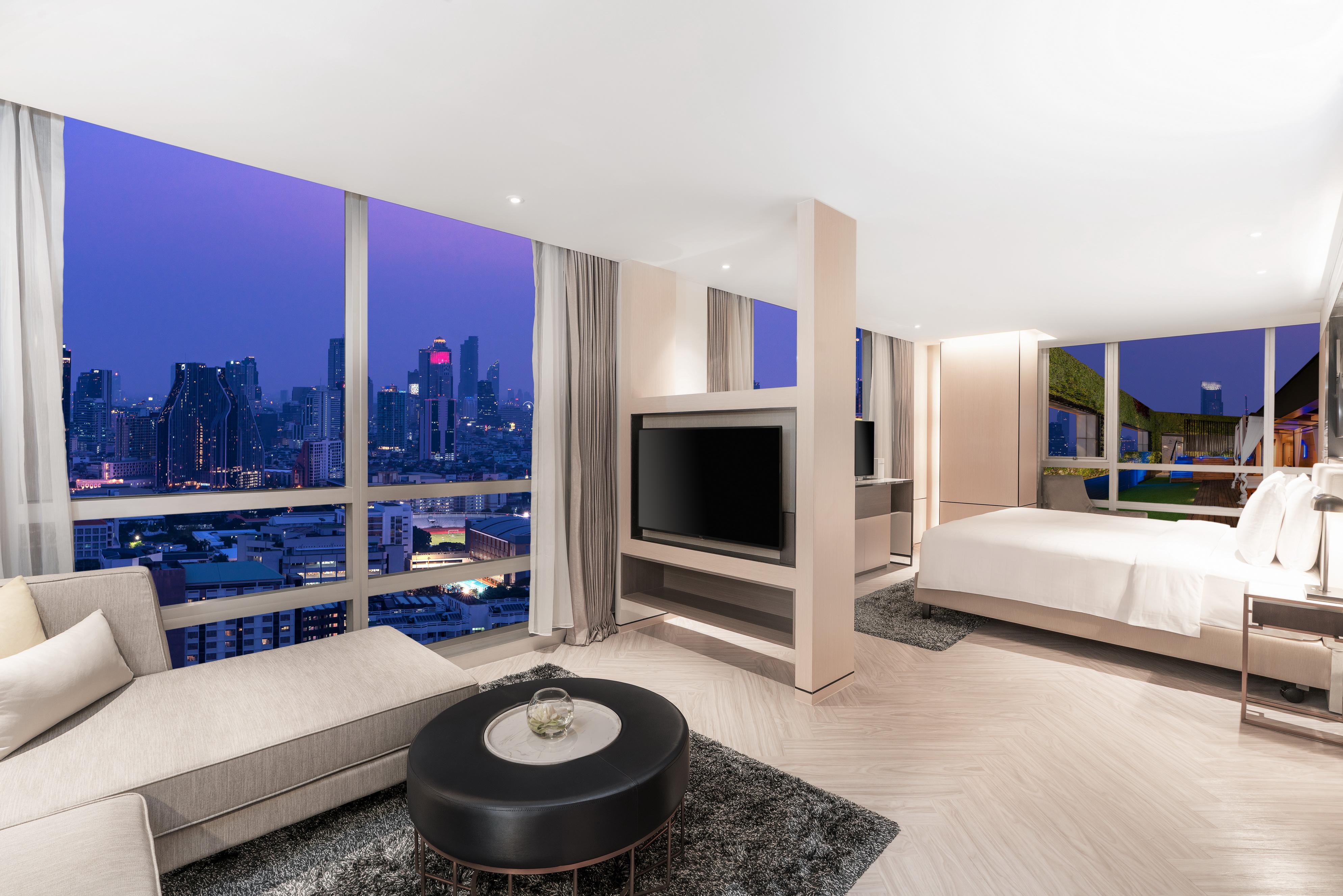 帕色哇公主酒店【SHA Extra Plus】 曼谷 外观 照片 The photo shows a modern hotel room with a stylish and contemporary design. There's a large window offering a panoramic view of a city skyline at dusk, with soft purple and blue hues in the sky. The room features a comfortable lounge area with a ligh