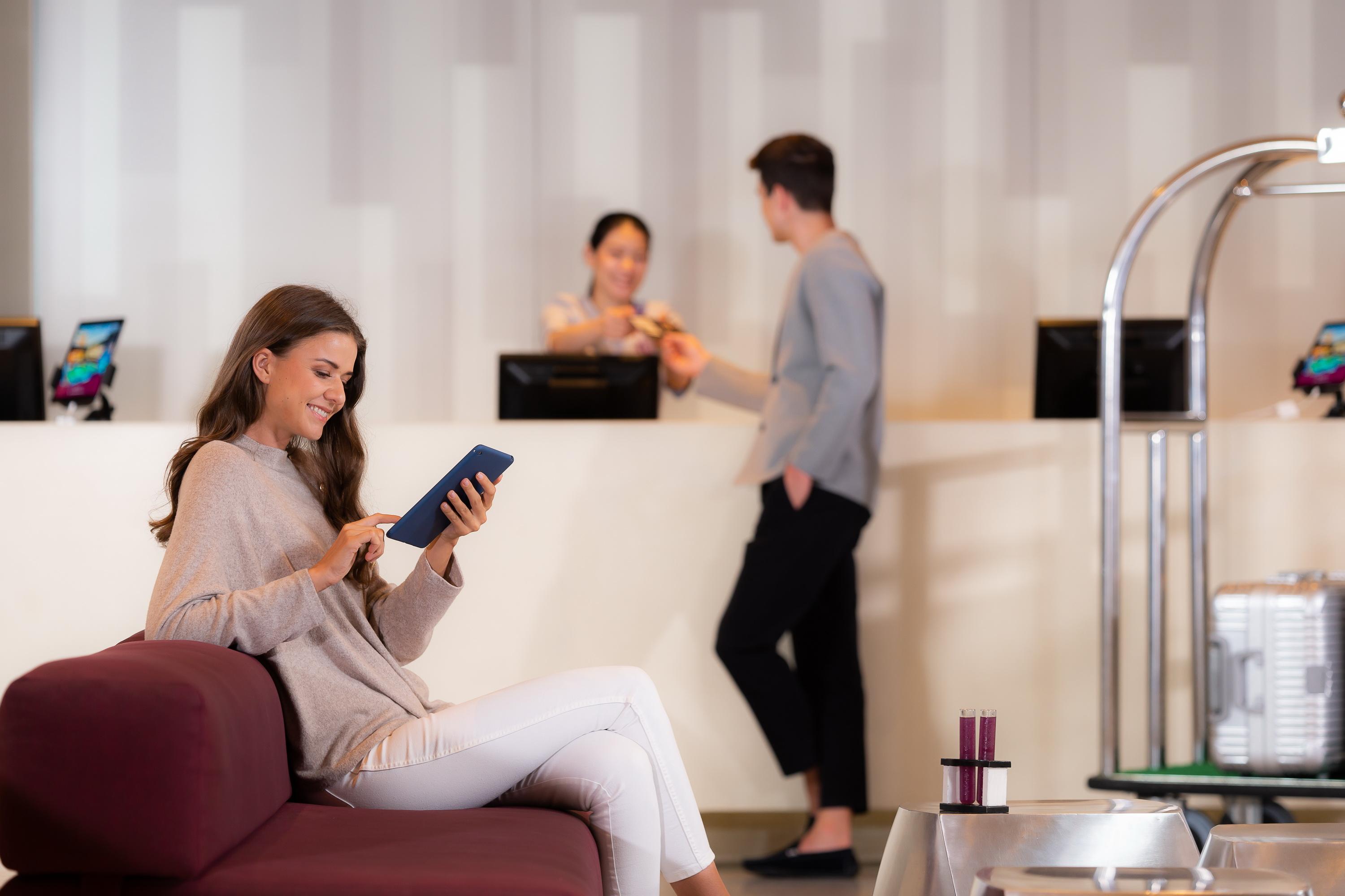 帕色哇公主酒店【SHA Extra Plus】 曼谷 外观 照片 The photo shows a modern reception area. In the foreground, a woman is sitting on a maroon sofa, engaged with a tablet. She is wearing a light sweater and white pants. In the background, two people are at the reception desk. One person is a staff mem