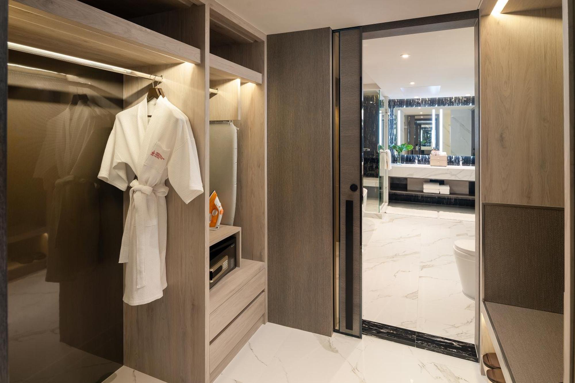 帕色哇公主酒店【SHA Extra Plus】 曼谷 外观 照片 The photo shows a modern bathroom area featuring sleek wooden cabinetry and marble flooring. There is a white bathrobe hanging on a hanger within an open closet space. The design is minimalist, with clean lines and a contemporary aesthetic. To the ri