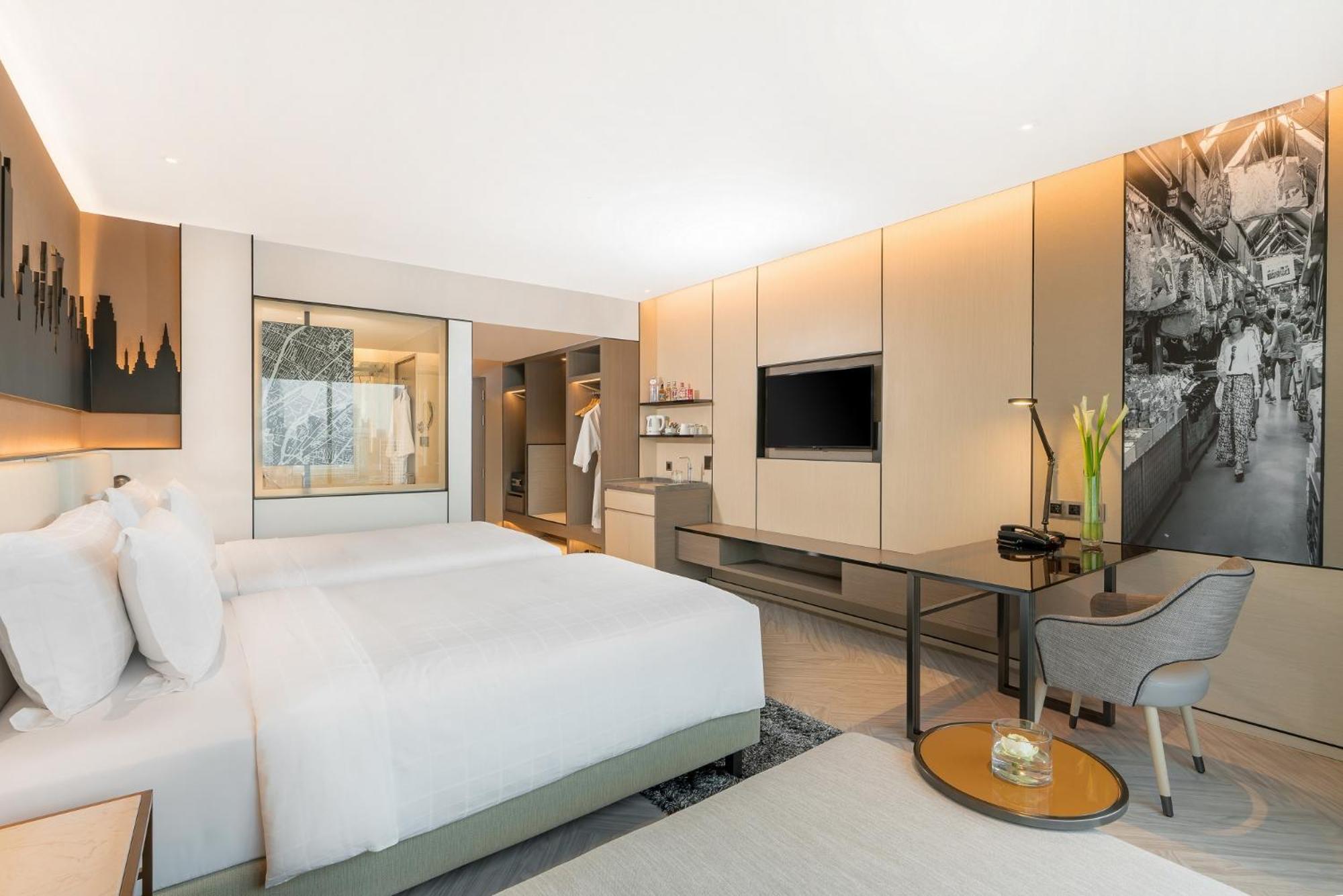 帕色哇公主酒店【SHA Extra Plus】 曼谷 客房 照片 The photo shows a modern hotel room featuring a minimalist design. There are two beds with white bedding, positioned against one wall. A stylish desk and chair are situated near a large window, which allows natural light to fill the space. A flat-scr