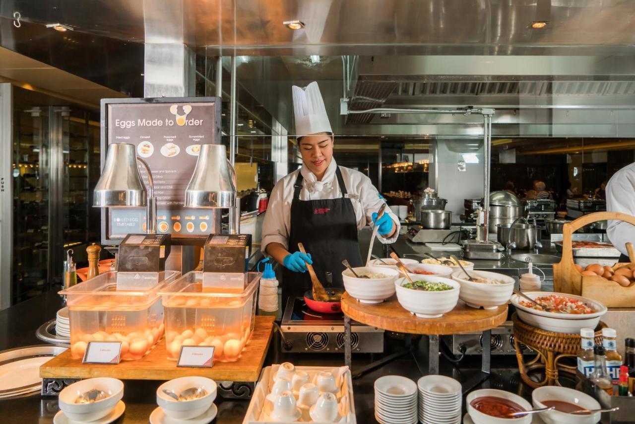 帕色哇公主酒店【SHA Extra Plus】 曼谷 外观 照片 The photo shows a chef working in a bustling kitchen environment. He is wearing a white chef's hat and an apron, along with blue gloves for hygiene. In front of him, there are several bowls and ingredients for preparing dishes. A display sign indicat